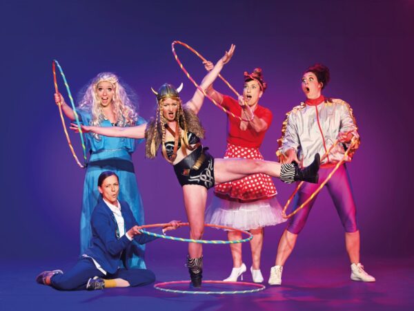 Auf dem Bild sind fünf Frauen vor einem violett-blauen Hintergrund zu sehen. Die Szene wirkt bunt, lebendig und humorvoll – fast wie ein Moment aus einer Zirkus- oder Comedy-Show. In der Mitte steht eine Frau in einem Wikinger-Kostüm: Sie trägt einen Helm mit Hörnern, ein Fellteil über den Schultern und hebt ein Bein hoch in die Luft, als würde sie gerade eine akrobatische Bewegung machen. Ihr Gesichtsausdruck ist energisch und leicht übertrieben, fast kämpferisch. Um sie herum stehen vier weitere Frauen, jede in einem ganz eigenen, auffälligen Kostüm: Links steht eine Frau mit langen, hellblonden, lockigen Haaren in einem fließenden blauen Kleid. Sie hält einen bunten Hula-Hoop-Reifen und schaut überrascht mit offenem Mund. Davor, am Boden sitzend, ist eine Frau in einem blauen Anzug. Sie hält ebenfalls einen Reifen und blickt konzentriert nach oben, als würde sie die Aktion unterstützen. Rechts neben der Wikingerin steht eine Frau in einem roten Kleid mit weißen Punkten (ähnlich einem Retro-Stil). Sie hebt die Arme und wirkt erstaunt oder begeistert. Ganz rechts steht eine Frau in einem silbrig glänzenden Outfit mit kurzen Hosen und Sneakers. Auch sie hält einen Reifen und schaut mit weit aufgerissenen Augen überrascht zur Mitte. Mehrere bunte Hula-Hoop-Reifen sind im Bild verteilt – einige werden gehalten, andere liegen am Boden. Die gesamte Gruppe wirkt wie in einer eingefrorenen, chaotisch-komischen Bewegung, voller Energie, Überraschung und Spielfreude. Insgesamt vermittelt das Bild eine Mischung aus Akrobatik, Clownerie und Theater – lebendig, witzig und ein bisschen verrückt.