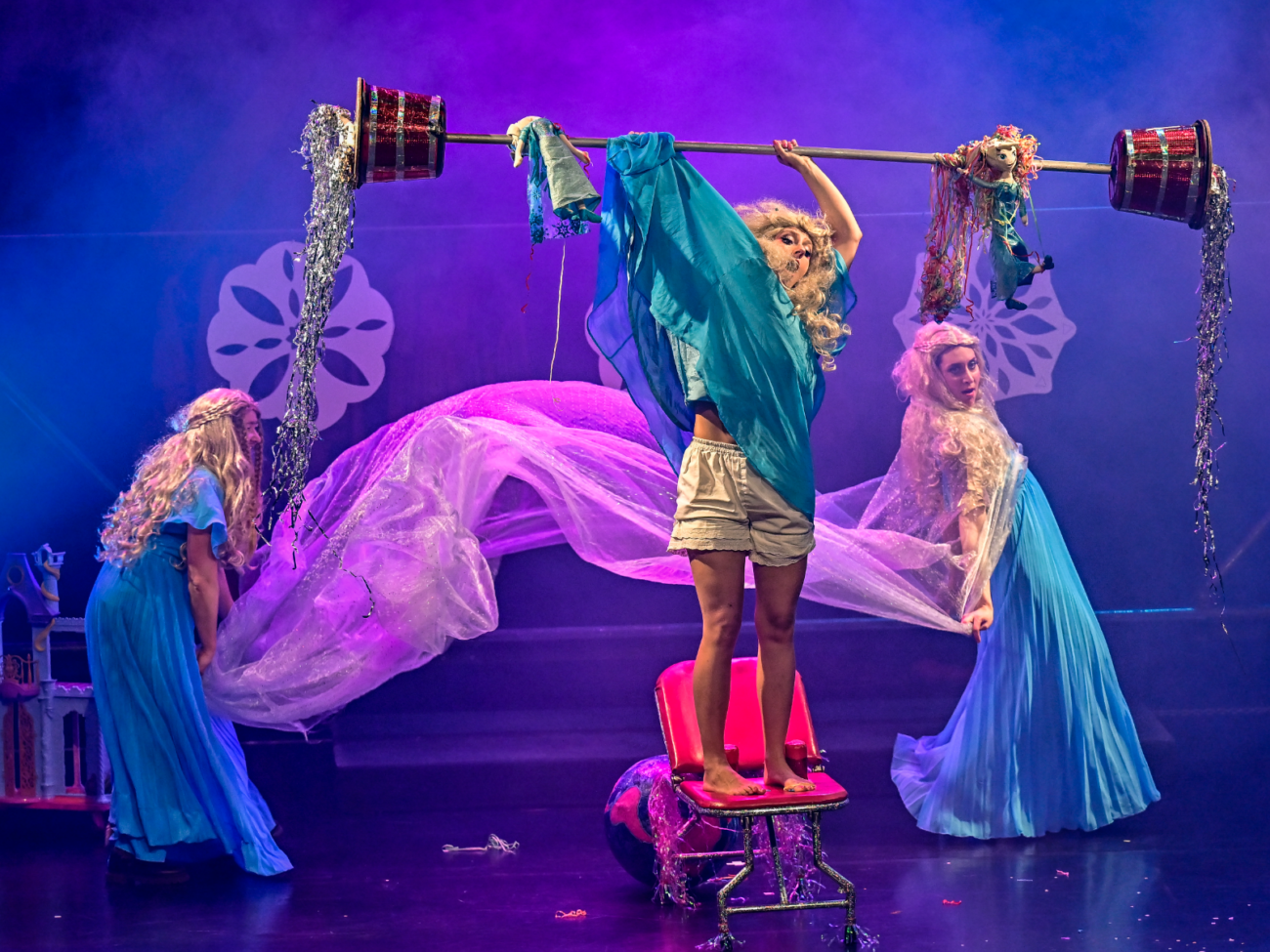 Das Bild zeigt eine farbenfrohe Bühnenszene mit drei Frauen, die wie in einer fantasievollen Theater- oder Zirkusaufführung inszeniert sind. Die Bühne ist in violett-blaues Licht getaucht, mit leichtem Nebel im Hintergrund. Im Mittelpunkt steht eine Frau auf einem Stuhl. Sie balanciert barfuß auf der Sitzfläche und hält über ihrem Kopf eine lange Stange, ähnlich wie eine Hantel. An beiden Enden dieser Stange hängen dekorative, zylinderförmige Behälter, aus denen silbernes Lametta herabfließt. Zusätzlich sind Stoffe und kleine Puppen an der Stange befestigt, die herunterhängen und der Szene etwas Verspieltes geben. Die Frau trägt ein lockeres, türkisfarbenes Oberteil und kurze helle Hosen. Ihr Gesichtsausdruck wirkt konzentriert und leicht angespannt, als würde sie sich auf das Gleichgewicht fokussieren. Links und rechts von ihr stehen zwei weitere Frauen in langen, fließenden, hellblauen Kleidern und mit langen, blonden Perücken. Sie halten ein großes, transparentes Tuch, das sich wie eine Welle durch die Luft zieht. Die Bewegung des Stoffes wirkt weich und elegant, fast wie Wasser oder Nebel. Der Hintergrund ist mit dekorativen, hellen Formen gestaltet, die an Blumen oder Ornamente erinnern. Auf dem Boden liegen kleine Requisiten und Glitzerreste, die die verspielte, leicht chaotische Atmosphäre verstärken. Insgesamt vermittelt die Szene eine Mischung aus Balancekunst, Theater und märchenhafter Fantasie – gleichzeitig zart, komisch und ein bisschen surreal.