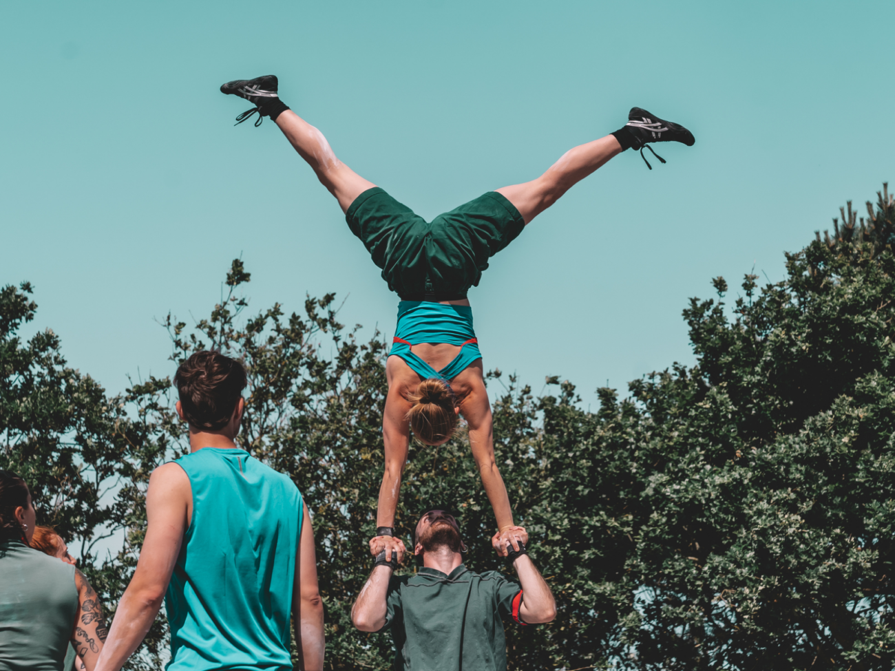 Das Bild zeigt eine akrobatische Szene im Freien vor einem blauen Himmel und grünen Bäumen. Im Mittelpunkt steht eine Frau, die einen Handstand macht – allerdings nicht auf dem Boden, sondern auf den Händen eines Mannes. Der Mann steht aufrecht und hält ihre Hände fest über seinem Kopf. Er trägt sie also komplett in die Höhe, während sie kopfüber balanciert. Ihr Körper ist gestreckt und kraftvoll: Die Arme sind gerade nach unten gerichtet (zu seinen Händen), der Oberkörper stabil, und die Beine weit auseinander gespreizt, fast wie ein großes „V“ in der Luft. Sie trägt sportliche Kleidung – ein kurzes Oberteil und Shorts – sowie Turnschuhe. Der Mann unter ihr schaut nach oben zu ihr und wirkt konzentriert und stabil, da er ihr gesamtes Gewicht trägt und gleichzeitig das Gleichgewicht halten muss. Links im Bild stehen weitere Personen, vermutlich Teil der Gruppe, die zuschauen oder bereit sind zu helfen. Sie tragen ebenfalls sportliche Kleidung. Die Szene vermittelt große körperliche Kontrolle, Kraft und Vertrauen zwischen den beiden: eine Partnerakrobatik-Figur, die gleichzeitig elegant und sehr anspruchsvoll wirkt.