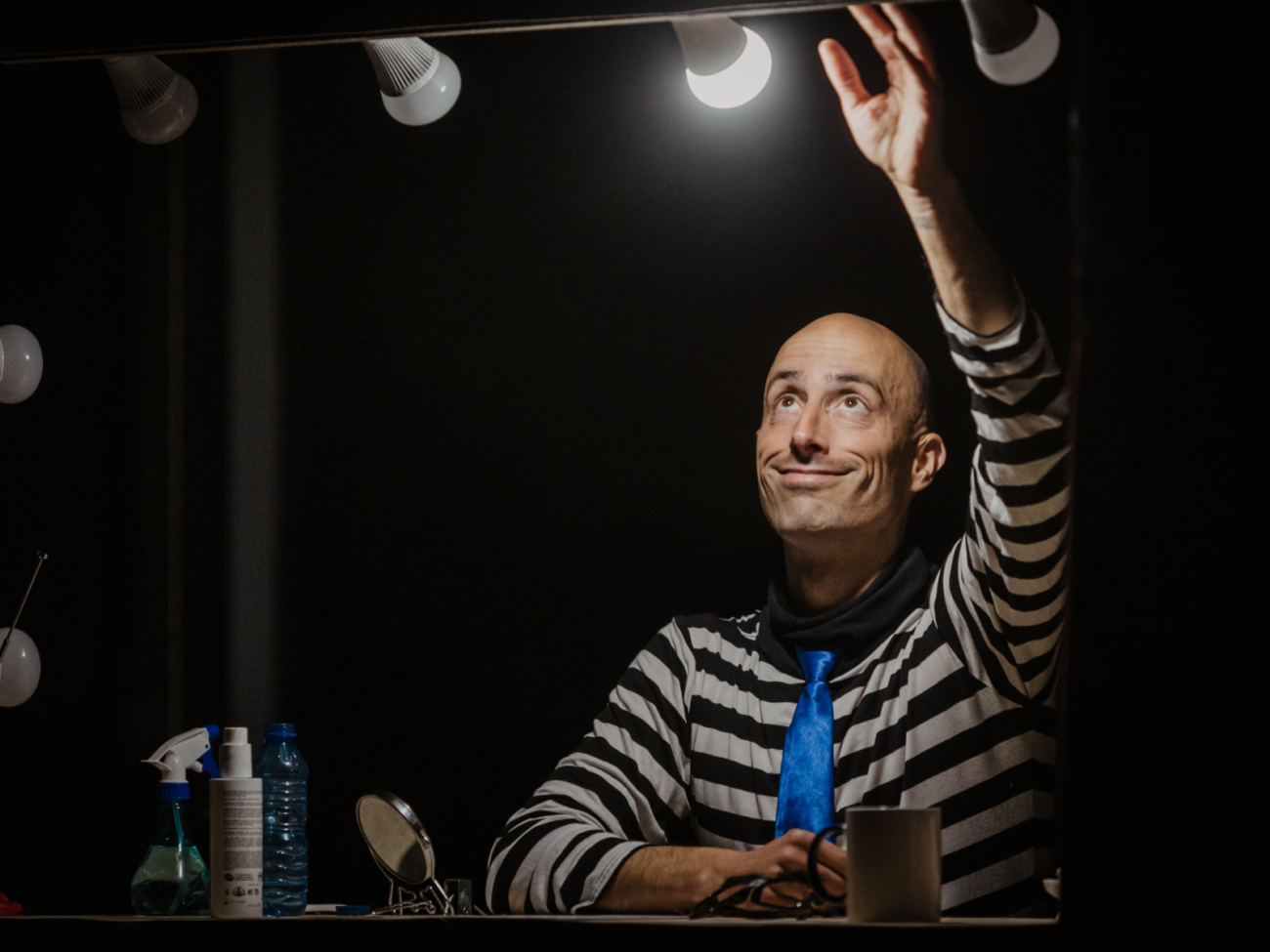 Das Bild zeigt einen Mann, der vor einem beleuchteten Schminkspiegel sitzt, wie in einer Theatergarderobe. Der Spiegel ist oben mit mehreren runden Glühbirnen versehen, die helles, warmes Licht abgeben. Der Hintergrund ist dunkel, sodass der Mann und sein Gesicht stark hervorgehoben werden. Der Mann ist kahlköpfig und trägt ein schwarz-weiß gestreiftes Oberteil, das an einen Clown oder eine Bühnenfigur erinnert. Dazu trägt er eine auffällig leuchtend blaue Krawatte, die einen starken Farbkontrast bildet. Er hebt einen Arm nach oben und streckt die Hand in Richtung einer der Glühbirnen aus, als würde er sie berühren oder nach ihr greifen. Dabei schaut er nach oben zu der Lampe, mit einem leicht verspielten, fast schelmischen Lächeln im Gesicht. Vor ihm auf dem Tisch stehen verschiedene Gegenstände: Flaschen, vermutlich für Schminke oder Reinigung, ein kleiner Spiegel und eine Tasse. Diese Dinge deuten darauf hin, dass er sich gerade vorbereitet oder zwischen zwei Auftritten befindet. Die Stimmung wirkt ruhig, konzentriert und zugleich ein wenig humorvoll – wie ein stiller, persönlicher Moment hinter der Bühne, in dem sich ein Künstler mit Licht, Ausdruck und seiner Rolle beschäftigt.