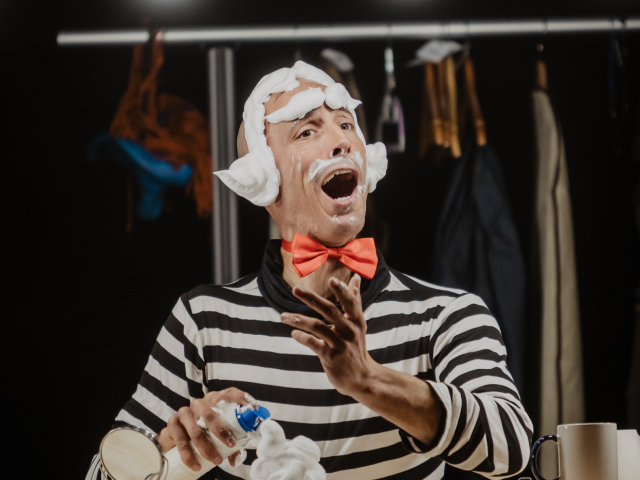 Das Bild zeigt einen Mann in einer Theatergarderobe, der sich gerade in eine Figur verwandelt – vermutlich eine clowneske Rolle. Er trägt ein schwarz-weiß gestreiftes Oberteil und eine auffällige rote Fliege um den Hals. Sein Kopf ist teilweise mit weißem Schaum bedeckt, der wie eine verspielte, übertriebene Frisur geformt ist – links und rechts wie kleine „Hörner“ oder Locken. Auch um seinen Mund und auf seinem Gesicht sind Reste des Schaums zu sehen. Sein Gesichtsausdruck ist sehr lebendig: Der Mund ist weit geöffnet, als würde er singen, rufen oder eine lustige Szene spielen. Die Augen sind nach oben gerichtet, und insgesamt wirkt seine Mimik übertrieben und theatralisch – typisch für Comedy oder Clownerie. Mit einer Hand hält er eine Dose oder Flasche, aus der der weiße Schaum kommt. Die andere Hand ist nach vorne ausgestreckt, als würde er gestikulieren oder das Publikum ansprechen. Im Hintergrund sieht man unscharf eine Kleiderstange mit Kostümen. Die Beleuchtung ist warm und konzentriert auf ihn, wodurch er klar im Mittelpunkt steht. Die Szene wirkt humorvoll, verspielt und ein bisschen chaotisch – ein Moment mitten in der Verwandlung oder bereits Teil einer komischen Darbietung.