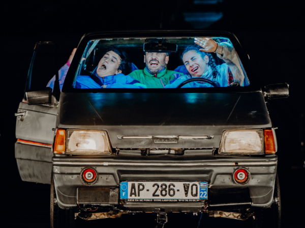 Das Bild zeigt die Front eines kleinen Autos direkt von vorne gesehen. Die Scheinwerfer sind eingeschaltet, und das Fahrzeug wirkt leicht abgenutzt. Durch die Windschutzscheibe sieht man drei Personen, die dicht nebeneinander sitzen: In der Mitte sitzt eine Person am Steuer. Sie schaut nach vorne, das Gesicht angespannt, mit zusammengekniffenen Augen und geöffnetem Mund, als würde sie sich anstrengen oder laut reagieren. Links daneben sitzt eine weitere Person, die den Kopf zur Seite geneigt hat und ebenfalls den Mund offen hat – ihr Ausdruck wirkt übertrieben, fast wie in einer komischen Szene. Rechts sitzt eine dritte Person, die eine Hand nach oben hebt, als wolle sie sich schützen oder etwas abwehren. Auch ihr Gesicht ist angespannt, mit zusammengekniffenen Augen. Alle drei tragen farbige, eher sportliche Kleidung in Blau- und Grüntönen. Die Szene wirkt wie ein eingefrorener Moment voller Chaos oder Aufregung – als würde das Auto gerade ruckeln, bremsen oder in eine turbulente Situation geraten sein. Durch die starke Beleuchtung und die übertriebenen Gesichtsausdrücke entsteht eine humorvolle, theatralische Wirkung, typisch für Comedy oder Clownerie auf der Bühne.