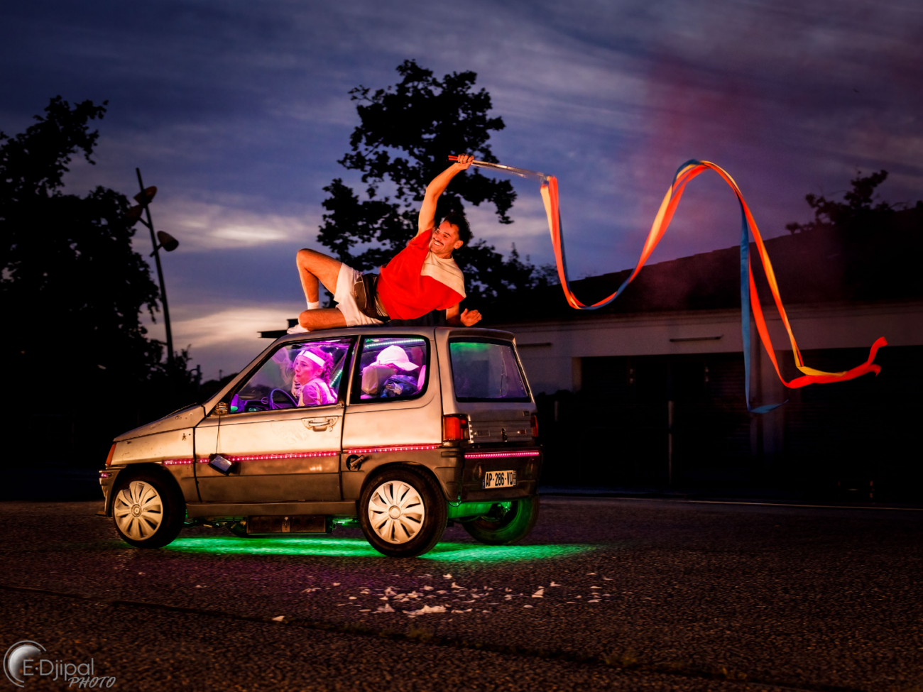 Das Bild zeigt eine lebendige, fast surreal wirkende Szene im Freien bei Dämmerung oder Nacht. Im Mittelpunkt steht ein kleines Auto. Unter dem Auto leuchtet grünes Licht, und auch im Innenraum sind bunte, violette und pinke Lichter zu sehen, die alles sehr theatralisch wirken lassen. Auf dem Dach des Autos liegt ein Mann in einer entspannten, halb liegenden Pose. Er stützt sich auf einen Arm und hält mit der anderen Hand einen langen Stab, an dem bunte Bänder befestigt sind. Diese Bänder wehen weit hinter ihm her und bilden geschwungene Linien in der Luft, als würden sie tanzen. Im Inneren des Autos sitzen zwei Personen. Sie wirken ebenfalls wie Teil einer Aufführung: Ihre Gesichter sind beleuchtet, und sie scheinen konzentriert oder überrascht zu reagieren. Das Auto selbst sieht etwas gebraucht aus, mit kleinen Gebrauchsspuren, was einen charmanten, improvisierten Eindruck vermittelt. Im Hintergrund erkennt man dunkle Silhouetten von Bäumen und Gebäuden gegen einen Abendhimmel mit blauen und violetten Farbtönen. Die gesamte Szene wirkt wie eine Mischung aus Straßentheater, Zirkus und Film – verspielt, bunt und voller Bewegung, als würde sich gerade eine fantasievolle Geschichte auf der Straße entfalten.
