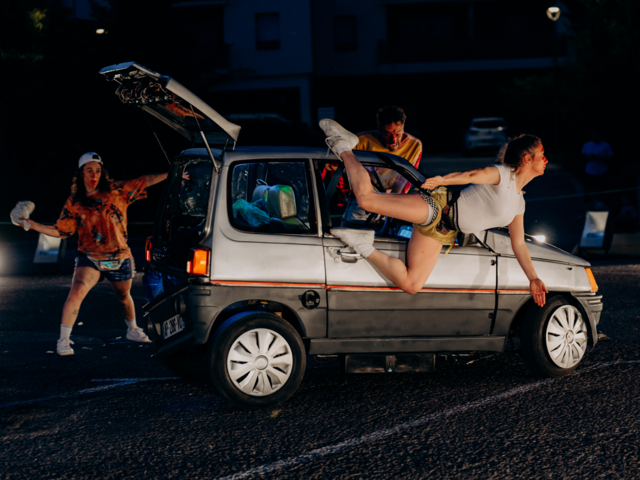 Das Bild zeigt eine dynamische, fast chaotisch wirkende Straßentheater-Szene bei Nacht. Im Mittelpunkt steht ein kleines Auto, das auf einer nassen Straße steht oder sich bewegt – der Boden glänzt im Licht, als hätte es geregnet. Die Beleuchtung ist dramatisch, mit starken Lichtquellen, die die Szene wie auf einer Bühne wirken lassen. Rechts am Auto hängt eine Frau halb aus dem Fenster. Ihr Körper ist waagerecht nach außen gestreckt, ein Bein ist nach oben angewinkelt, das andere nach hinten. Sie hält sich mit den Händen am Auto fest und wirkt, als würde sie gerade aus dem Fahrzeug „herausfliegen“. Ihr Gesicht ist angespannt und nach vorne gerichtet, als wäre sie mitten in einer actionreichen Bewegung. Im Auto selbst ist eine weitere Person zu sehen, die sich nach vorne beugt und ebenfalls sehr intensiv reagiert – mit offenem Mund und starkem Ausdruck, als wäre etwas Dramatisches passiert. Links hinter dem Auto steht eine dritte Person auf der Straße. Sie trägt auffällige, bunte Kleidung und hält etwas Weißes in der Hand. Ihre Arme sind ausgebreitet, und sie wirkt überrascht oder erschrocken, als würde sie auf die Szene reagieren. Die Heckklappe des Autos ist geöffnet, was den Eindruck von Unordnung oder Hektik verstärkt. Insgesamt wirkt das Bild wie ein eingefrorener Moment aus einer wilden, komischen Verfolgungs- oder Actionszene – voller Bewegung, Spannung und übertriebener Körpersprache, typisch für modernes Straßentheater oder Comedy.