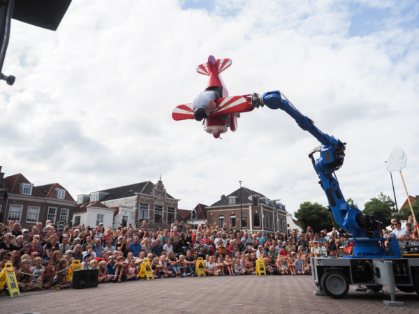 Das Bild zeigt eine große Straßentheater-Szene auf einem Platz in einer Stadt, bei Tageslicht. Im Vordergrund steht ein großer, blauer Roboterarm – ähnlich wie in einer Fabrik. Dieser Arm hebt ein kleines rotes Flugzeug mit weißen Streifen hoch in die Luft. Das Flugzeug wirkt wie ein fantasievolles Requisit. In diesem Flugzeug steckt eine Person kopfüber drin: Der Oberkörper hängt nach unten aus dem Flugzeug heraus, während die Beine im Inneren verschwinden. Es sieht so aus, als würde die Person gerade aus dem Flugzeug „herausfallen“ oder darin feststecken – eine humorvolle, akrobatische Darstellung. Unterhalb dieser Szene sitzt und steht eine große Menschenmenge. Viele Zuschauerinnen und Zuschauer – darunter auch Kinder – schauen nach oben und verfolgen gespannt das Geschehen. Einige sitzen auf dem Boden in der ersten Reihe, andere stehen dahinter. Im Hintergrund sind Häuser mit Ziegeldächern und eine helle, leicht bewölkte Himmelskulisse zu sehen. Die gesamte Szene wirkt wie eine spektakuläre, spielerische Performance: eine Mischung aus Technik, Akrobatik und Humor, die das Publikum zum Staunen und Lachen bringt.