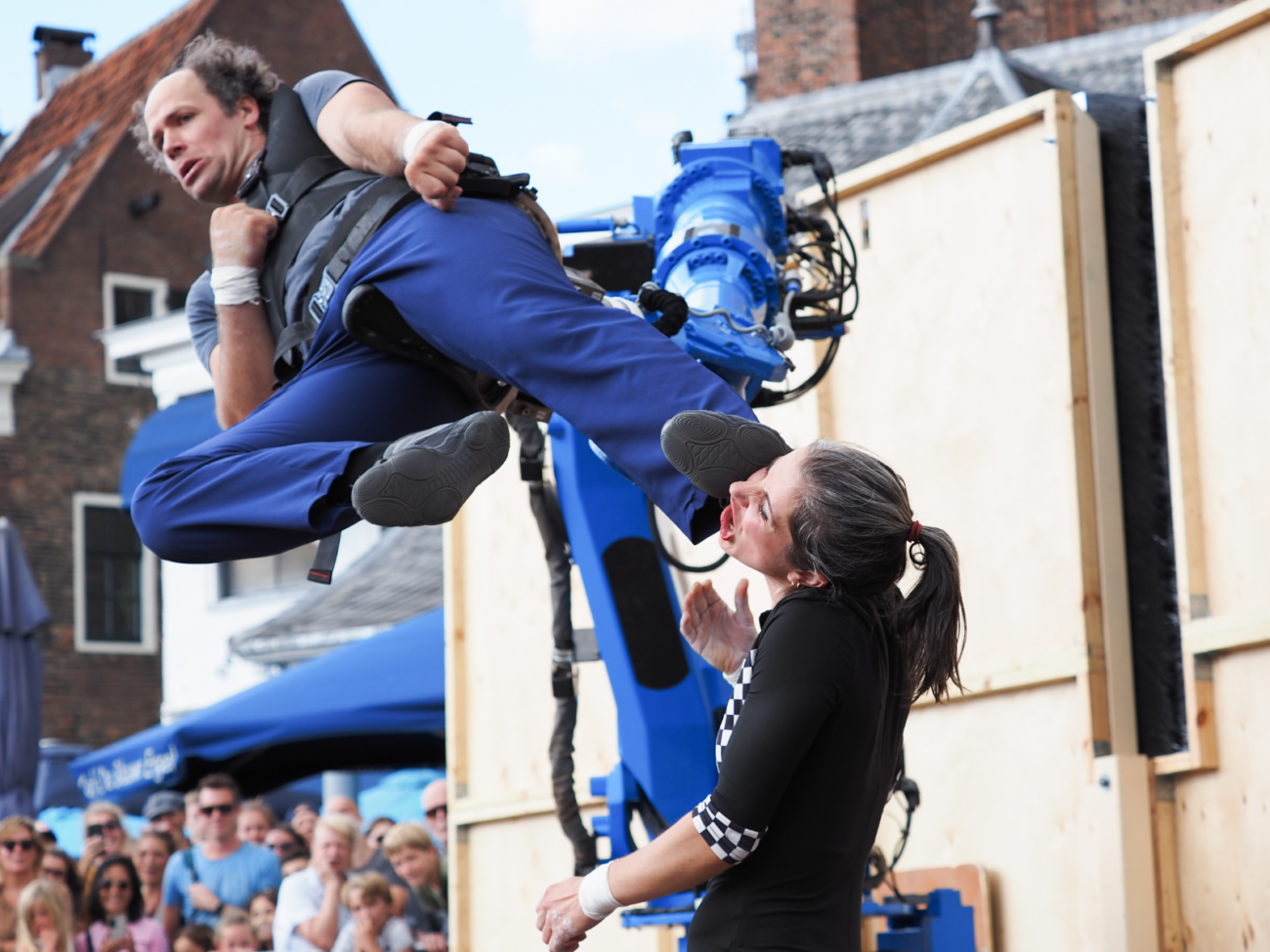Das Bild zeigt eine dynamische Straßentheater und Akrobatikszene vor Publikum. Im Mittelpunkt ist ein Mann in blauer Kleidung, der in der Luft hängt oder gehalten wird – sein Körper ist angewinkelt, als wäre er gerade in einer Bewegung eingefroren. Sein Gesicht wirkt angespannt und konzentriert. Er trägt eine Art Gurt oder Geschirr, was darauf hindeutet, dass er an einer Konstruktion befestigt ist. Unter ihm steht eine Frau mit dunklen Haaren, die nach oben schaut. Ihr Gesicht ist sehr ausdrucksstark – sie verzieht den Mund und wirkt überrascht oder erschrocken, fast so, als würde sie gleich getroffen werden. Tatsächlich ist der Fuß des Mannes sehr nah an ihrem Gesicht, was die Szene komisch und spannend zugleich macht. Im Hintergrund sieht man einen großen blauen Roboterarm, der vermutlich die Bewegung oder Position des Mannes steuert. Dahinter stehen viele Zuschauerinnen und Zuschauer, die die Szene aufmerksam beobachten. Die Situation wirkt wie ein eingefrorener Moment aus einer humorvollen, leicht übertriebenen Aktion: eine Mischung aus Technik, Akrobatik und Comedy, bei der Nähe, Timing und Reaktion eine wichtige Rolle spielen.