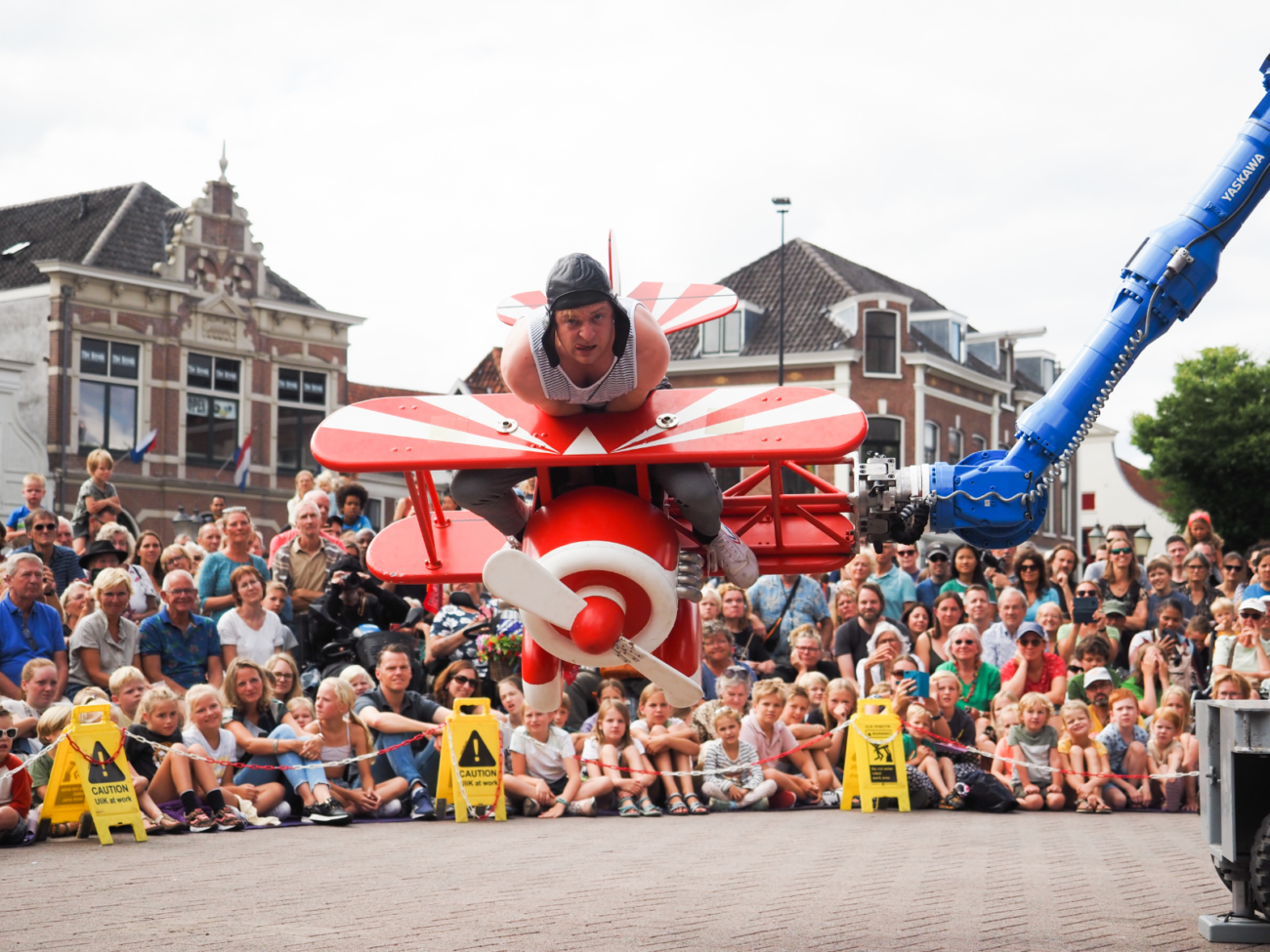 Das Bild zeigt eine Straßentheater-Szene auf einem Platz, aufgenommen aus der Perspektive des Publikums. Im Mittelpunkt schwebt ein kleines, rotes Flugzeug mit weißen Streifen direkt auf die Betrachter zu. Darauf liegt eine Person bäuchlings oben drauf, als würde sie selbst das Flugzeug steuern. Sie trägt eine Kappe und schaut nach vorne, mit einem konzentrierten, leicht angespannten Gesichtsausdruck. Das Flugzeug wirkt wie ein fantasievolles Bühnenobjekt und wird seitlich von einem großen blauen Roboterarm gehalten, der rechts im Bild zu sehen ist. Dadurch scheint das Flugzeug tatsächlich durch die Luft zu fliegen. Im Hintergrund sitzt und steht eine große Menschenmenge – viele Erwachsene und Kinder – dicht beieinander. Die Kinder sitzen vorne auf dem Boden, die Erwachsenen stehen dahinter. Alle schauen gespannt auf die Szene. Einige halten ihre Hände im Schoß, andere lehnen sich leicht nach vorne, um besser zu sehen. Zwischen Publikum und Bühne stehen gelbe Warnschilder und eine Absperrung, die den Aufführungsbereich markieren. Hinter den Menschen sieht man Häuser mit Ziegeldächern und einen hellen Himmel. Die Szene vermittelt den Eindruck eines spielerischen „Flugabenteuers“ mitten in der Stadt – eine Mischung aus Technik, Theater und Humor, die das Publikum direkt einbezieht und zum Staunen bringt.