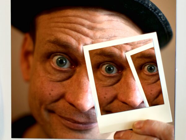 Zu sehen ist eine Nahaufnahme eines Mannes mit heller Haut, der direkt in die Kamera blickt. Er trägt einen schwarzen Hut und hat weit geöffnete, auffällige Augen, die dem Bild einen leicht überraschten oder spielerischen Ausdruck geben. Sein Gesicht ist sehr nah herangezoomt, sodass Stirn, Augen, Nase und Mund gut sichtbar sind. In seiner Hand hält er ein weißes, polaroidartiges Bild mit mehreren rechteckigen Ausschnitten. Diese Rahmen sind so vor sein Gesicht gehalten, dass Teile seines Gesichts – vor allem ein Auge – mehrfach innerhalb der Ausschnitte erscheinen. Dadurch entsteht ein visueller Effekt, bei dem sich sein Blick wiederholt und leicht verzerrt wirkt. Der Hintergrund ist unscharf und warm gehalten, sodass der Fokus ganz auf dem Gesicht und dem ungewöhnlichen Bildeffekt liegt. Insgesamt wirkt das Bild kreativ und verspielt, fast wie ein kleines optisches Experiment mit Perspektive und Wiederholung.